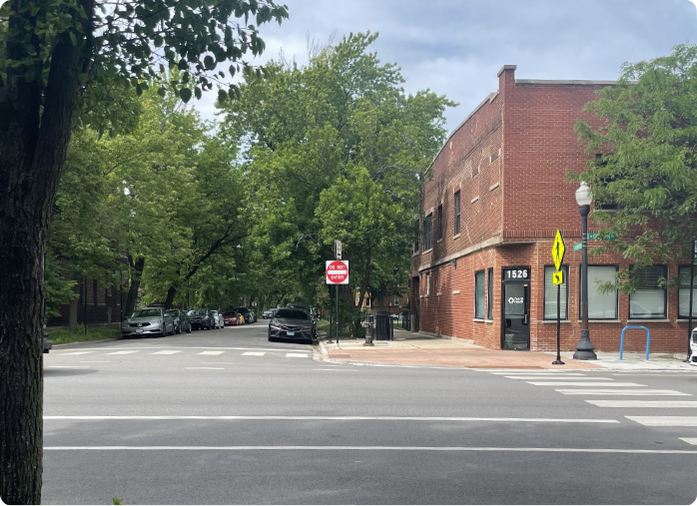 Urban street corner with a brick building, crosswalk, and tree-lined sidewalk.