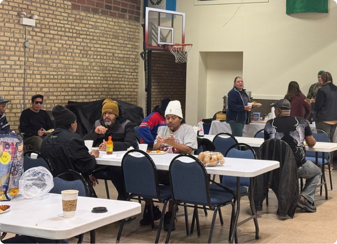 People seated at folding tables in a gym eating meals during a communal feed in a recreation hall