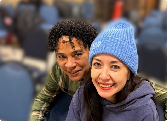 Two smiling women pose for a selfie in a crowded indoor setting; one wears a blue knit beanie and hoodie.