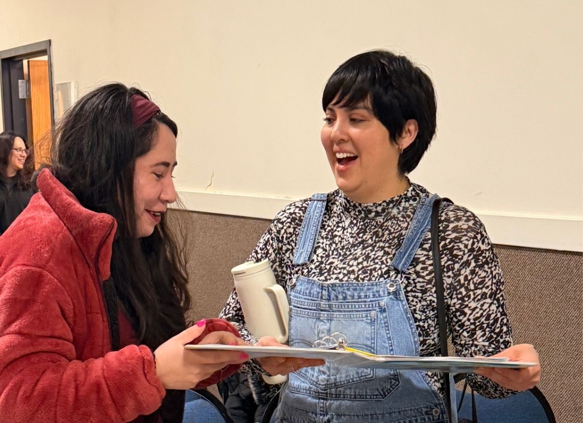 Two women chatting in a hallway; one in a red fleece jacket shows a clipboard to the other in denim overalls, both smiling with a white mug on the clipboard.